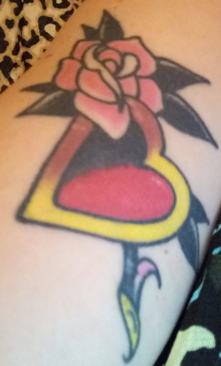 Aubrey shows off a tattoo of a pink rose and a red heart with gold rim. It is on her arm.