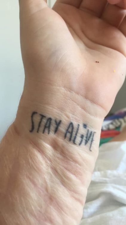 Hollie shows the viewer a tattoo reading "Stay alive." A semicolon takes the place of the 'i.' It is located on her wrist.