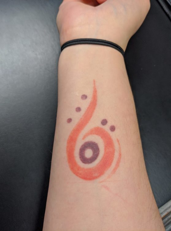 Cassie shows the viewer a tattoo of a ball of fire. It is located on her arm and is orange and red.