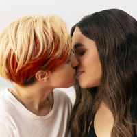 A transmasculine gender-nonconforming person and transfeminine non-binary person kissing.