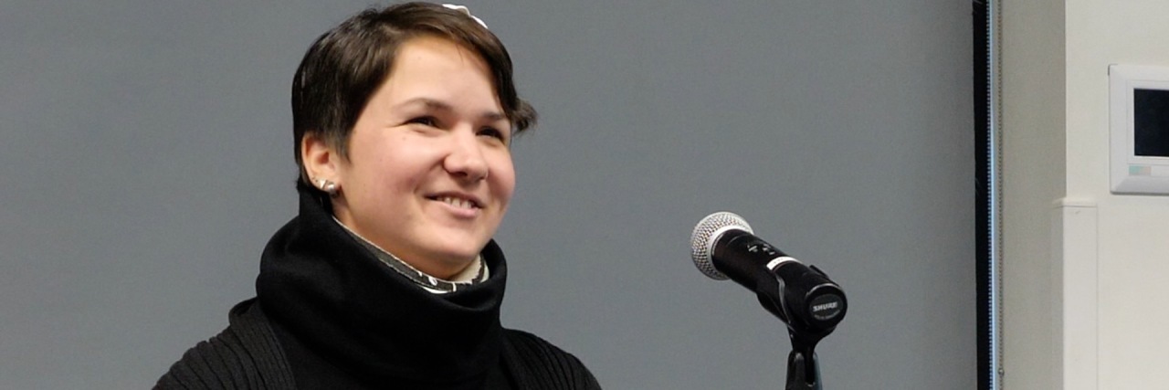 Karina, a woman with short brown hair sits at a podium and speaks to an audience. She smiles.