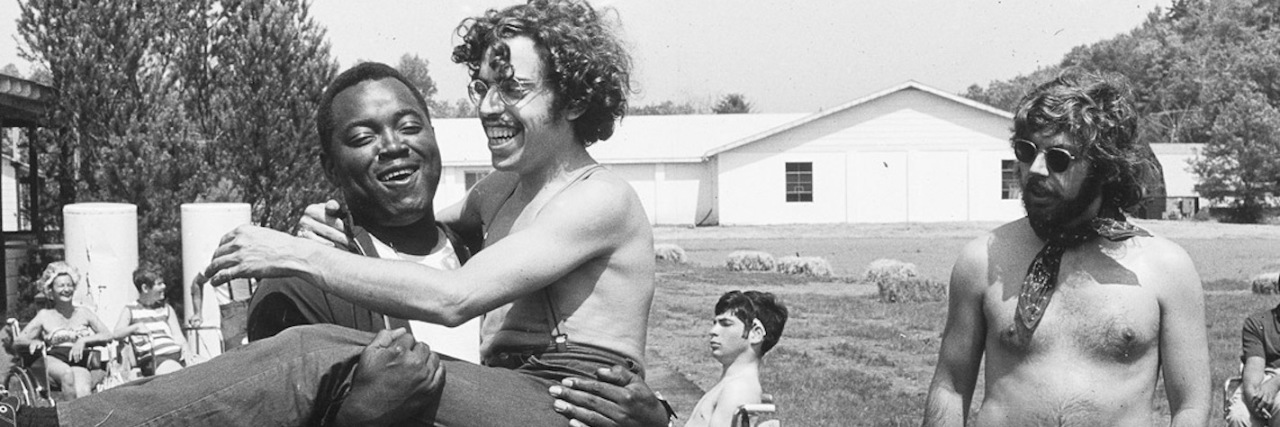 Netflix Documentary 'Crip Camp' Captures Disability Rights Movement Still image from the documentary Crip Camp showing a young black man carrying a young white man while another white man with long hair and a beard watches