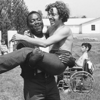 Still image from the documentary Crip Camp showing a young black man carrying a young white man while another white man with long hair and a beard watches