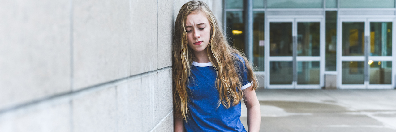 A teenage girl with long blonde hair leaning against a wall and looking down