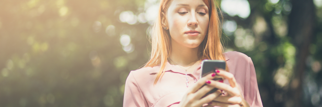 photo of young college student with phone in hands and sunlight on her face, trees behind her