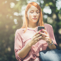 photo of young college student with phone in hands and sunlight on her face, trees behind her