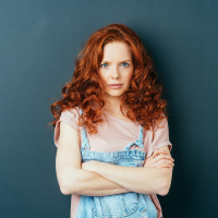 woman with curly red hair standing with arms folded staring at camera