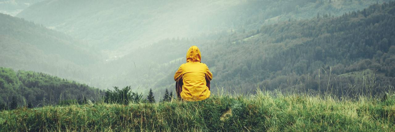 Learning to Appreciate Scars on Your Body Because of Illness person sitting in the grass, facing the mountains. sun shining through clouds