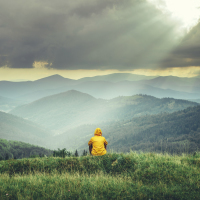 person sitting in the grass, facing the mountains. sun shining through clouds