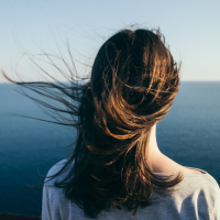 Woman with dark hair stands on a top cliff over blue sea