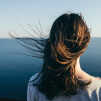 Woman with dark hair stands looking out over a blue sea view in the wind.
