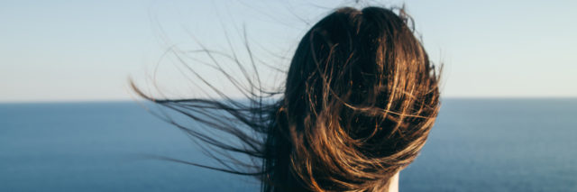 Woman with dark hair stands looking out over a blue sea view in the wind.