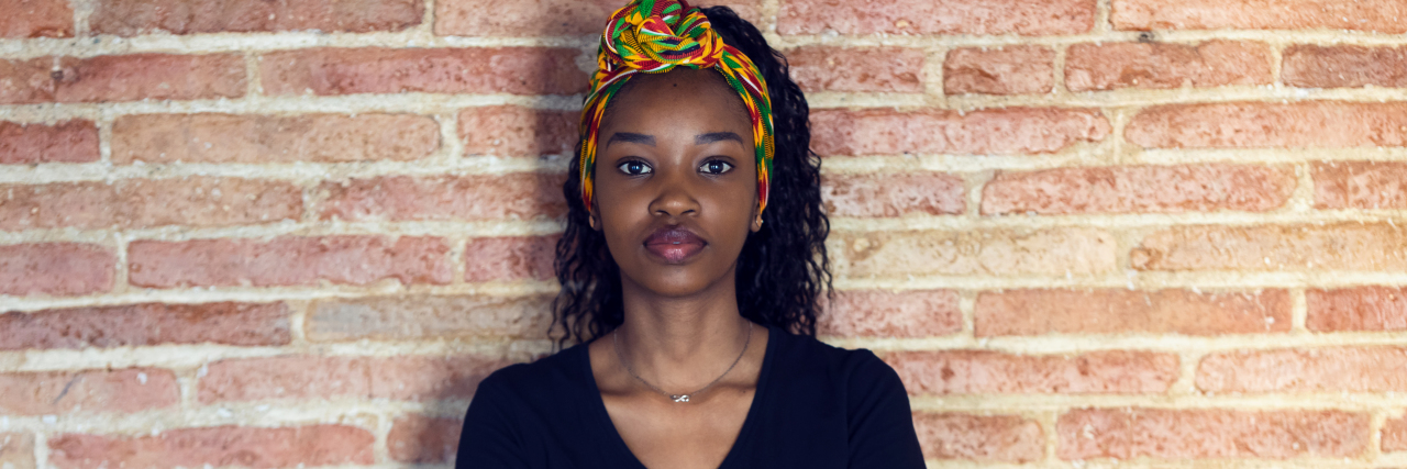 young black woman with a v neck shirt standing in front of a brick wall with her arms crossed and a straight face