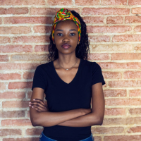 young black woman with a v neck shirt standing in front of a brick wall with her arms crossed and a straight face