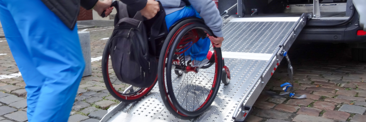 Assistant helping disabled person in wheelchair with transport using accessible van ramp.