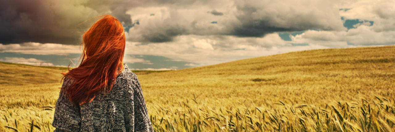 young woman standing in a wheat field, facing the sky and clouds