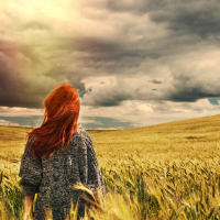 young woman standing in a wheat field, facing the sky and clouds