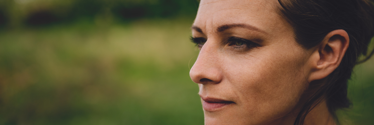 My Autoimmune Disease Makes Me Constantly Feel Guilty for Everything close up of a woman with brown hair looking to the side with a blurred green background of a field behind her
