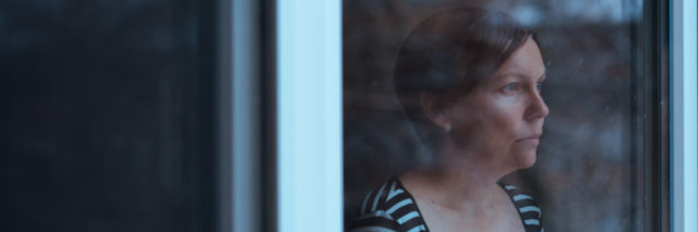 woman with short red hair in a striped shirt looking out the window solemnly