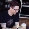photo of man wearing glasses and with arm tattoos sitting at table in cafe with coffee cup