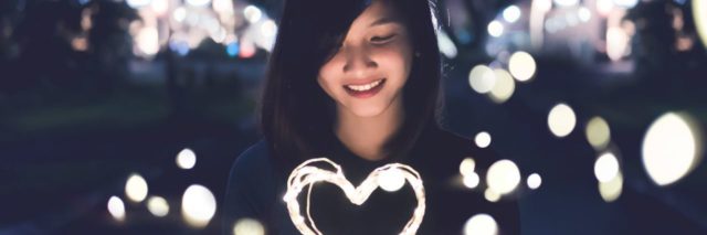 photo of woman looking down at a heart made out of light in her hands, smiling and surrounded by other lights