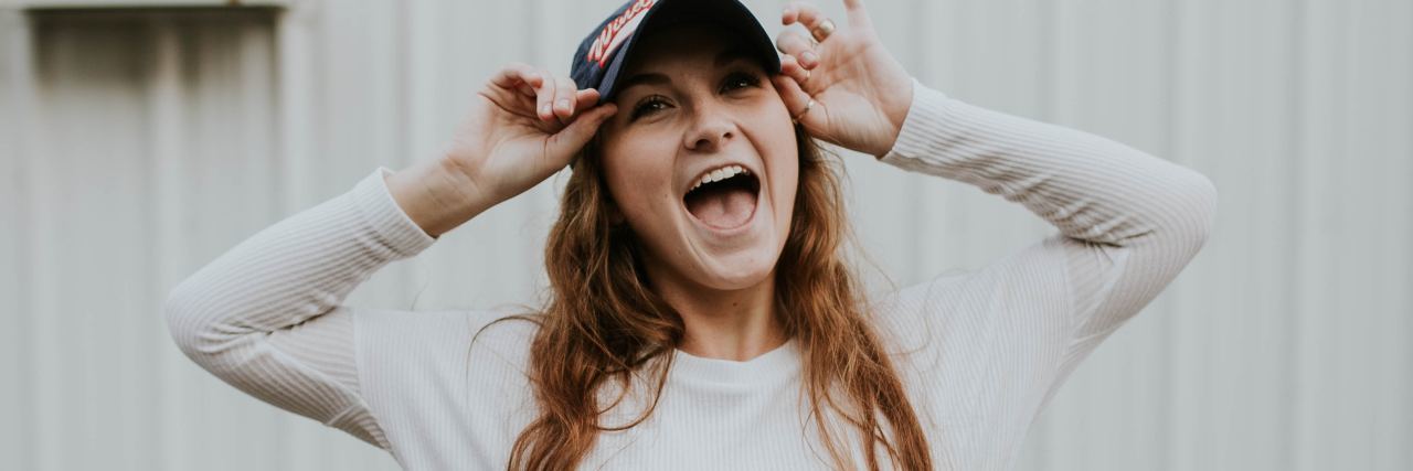 photo of happy young woman posing and laughing for camera with hands raised to baseball cap