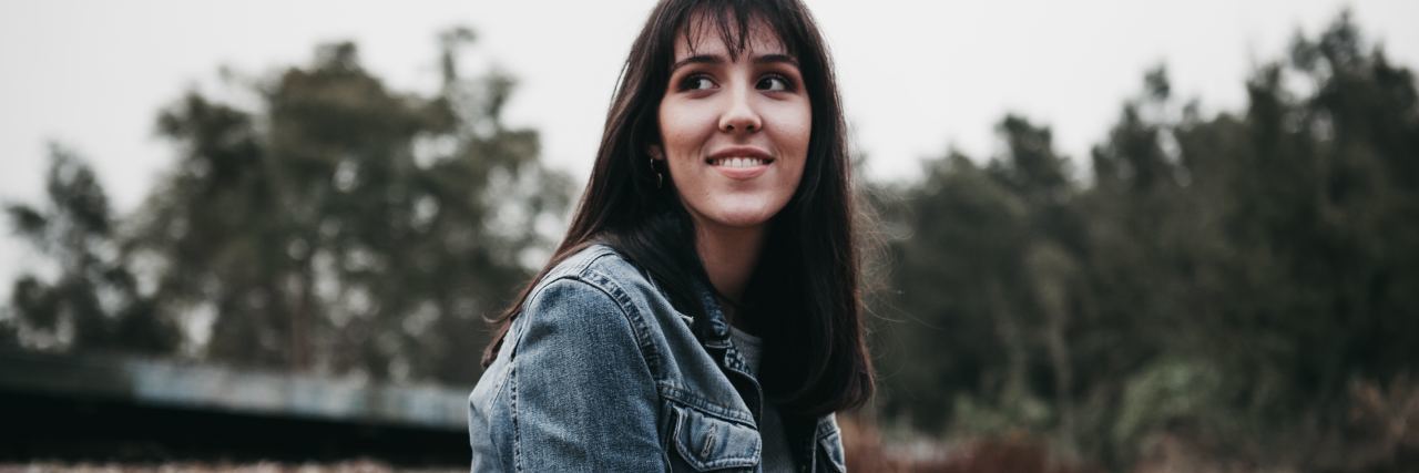 Learning to Take Care of My Health Instead of What Is Expected of Me photo of woman with dark hair looking back toward someone and smiling, with trees behind her