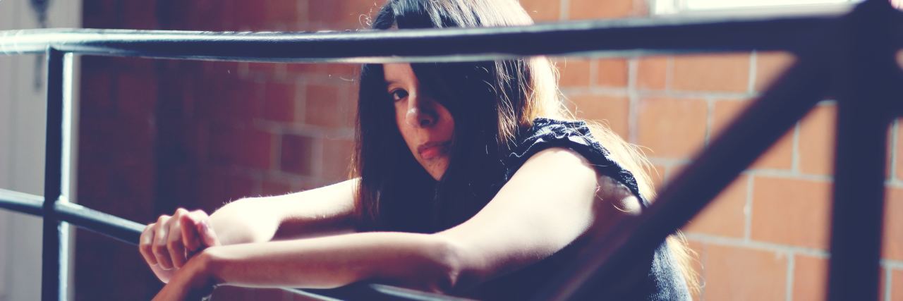 Why I Finally Felt Able to Tell My Therapist About My Self-Harm photo of young woman leaning on bars looking into camera with serious expression, a brick wall behind her