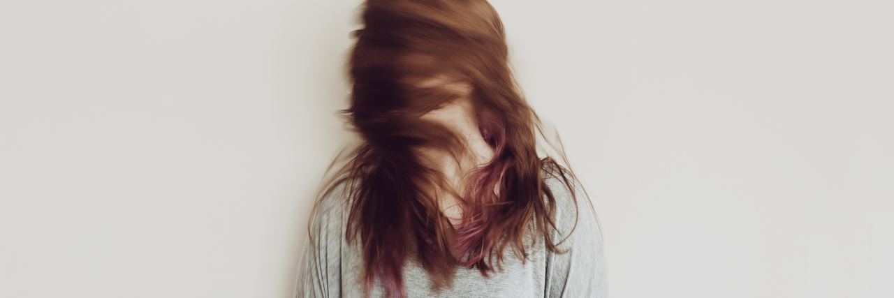 photo of woman standing against plain white wall, turning head fast so that her head is blurred and her hair covers her face
