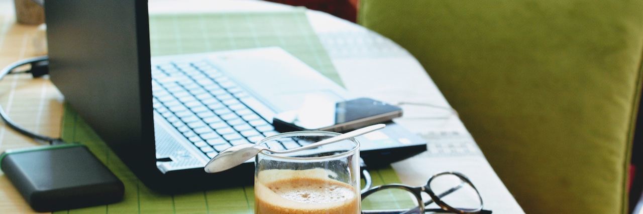 Remote Jobs for People With a Disability or Chronic Illness Computer on a desk with a pair of classes and cup of coffee on a wooden coaster nearby