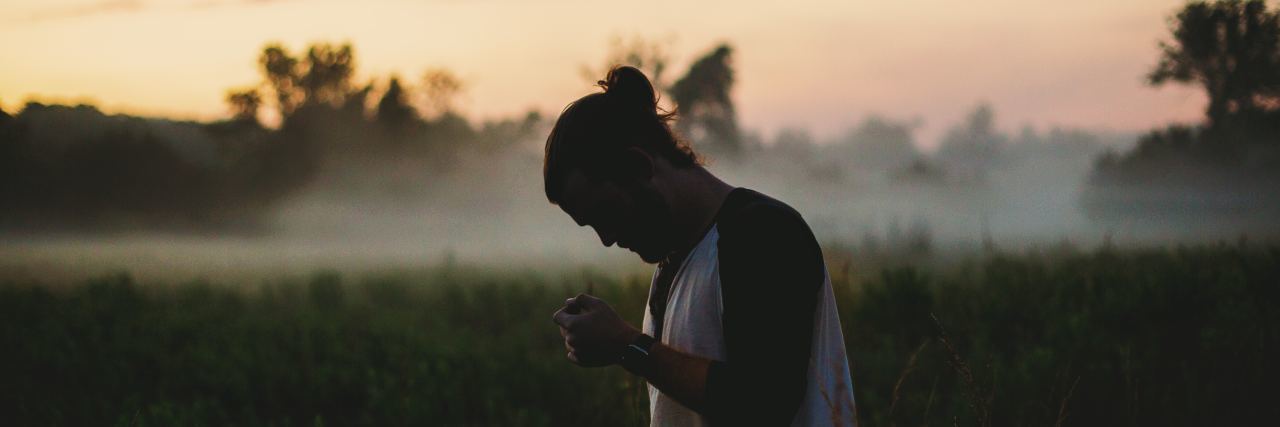 man with hair in a bun wearing a white shirt out in a field looking down with the sunset behind him