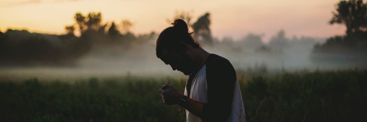 Please Understand These 5 Things About My Social Anxiety man with hair in a bun wearing a white shirt out in a field looking down with the sunset behind him