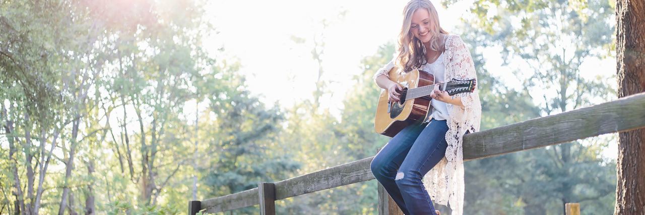 How Playing Guitar Saves Me From My Mental Illness photo of woman sitting on fence playing guitar in forest or park