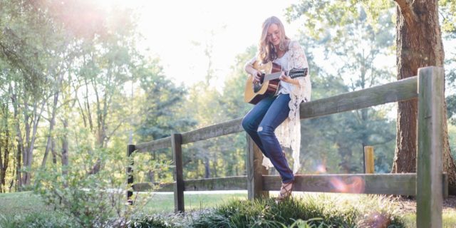 photo of woman sitting on fence playing guitar in forest or park