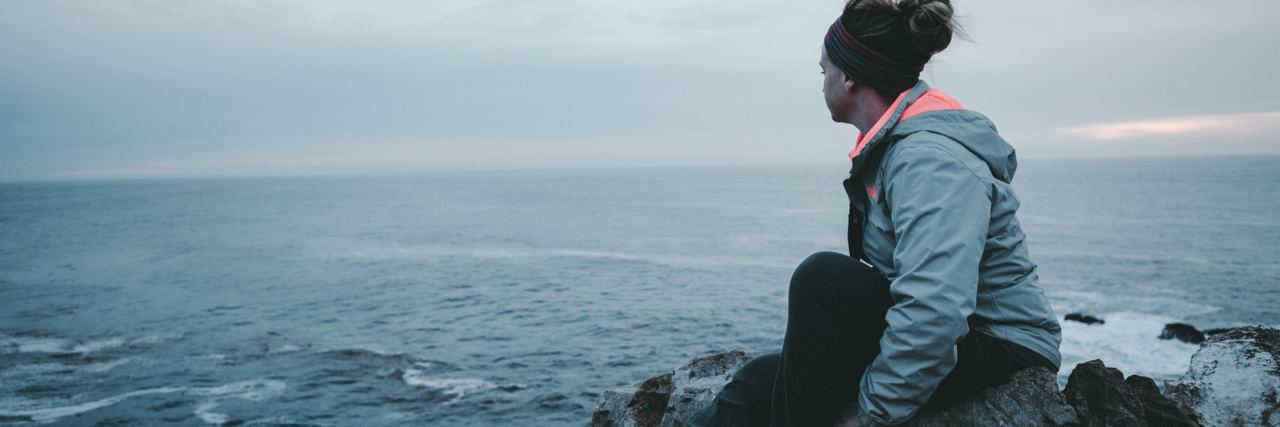 photo of woman sitting on rock by beach watching waves in distance