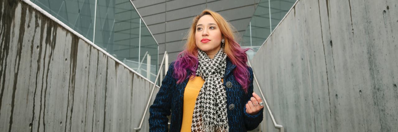 hispanic woman with blonde and purple hair wearing a yellow shirt and jean jacket and scarf walking down a hall looking ahead confidently