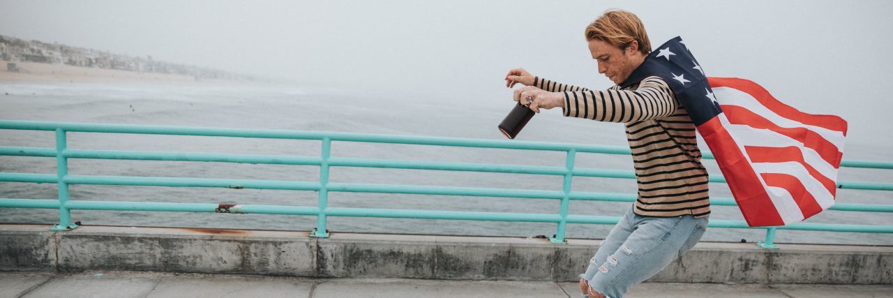 Living in 2020 With No Fear, Despite Depression and Anxiety young man with ripped jeans on the edge of a longboard with an American flag as a cape, looking down smiling on a sidewalk