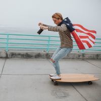 young man with ripped jeans on the edge of a longboard with an American flag as a cape, looking down smiling on a sidewalk