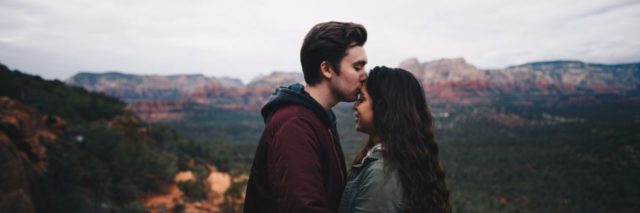 couple on top of a mountain, hugging each other with the boyfriend kissing the top of his girlfriend's head