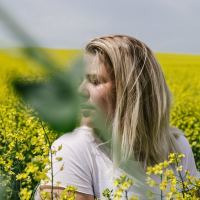 photo of blonde woman sitting in field of yellow flowers, looking away from camera, with a large leaf covering her face