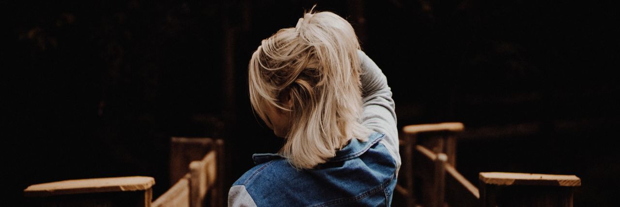How the DBT 'PLEASE' Skill Helps Me Practice Self-Care With Depression woman on a wooden bridge in the dark with her back facing the camera, holding her blonde hair up in her hand