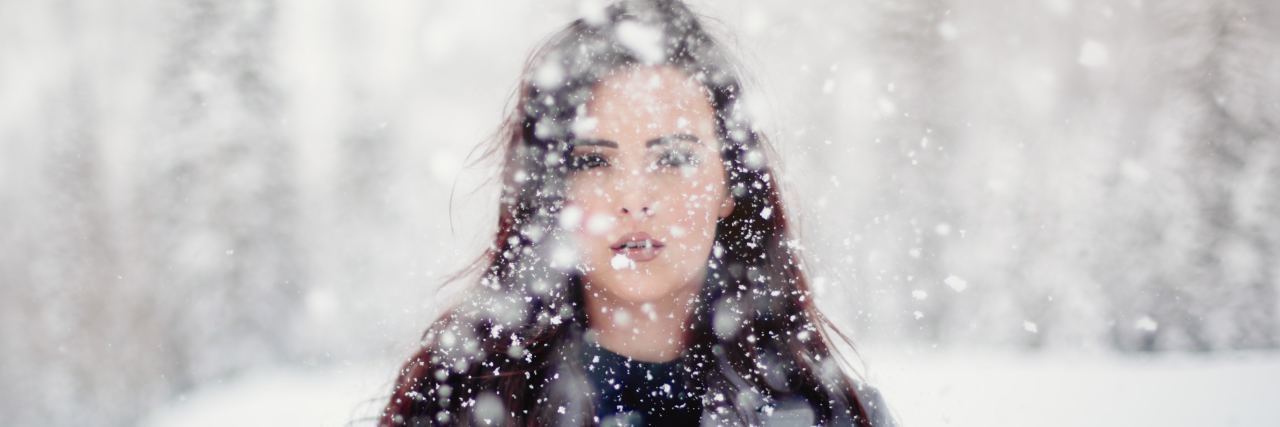 woman in a grey jacket with long brown hair staring at camera as snow falls down around her