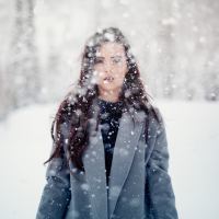 woman in a grey jacket with long brown hair staring at camera as snow falls down around her
