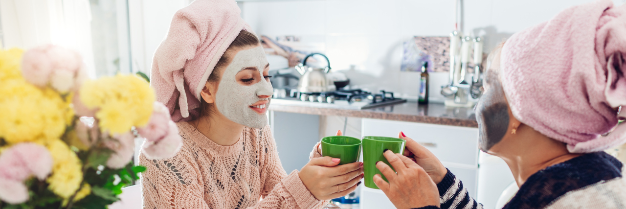 a mom and her adult daughter sipping from coffee cups in a kitchen with facial masks on