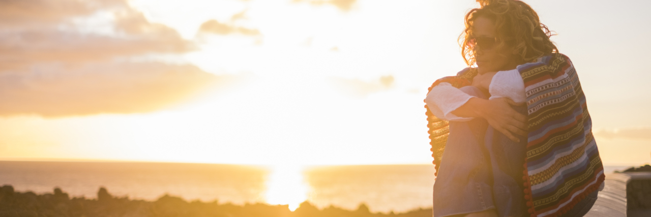 Why It's Important for Others With Ehlers-Danlos Syndrome to Speak Up a woman with curly hair is sitting outside on a wall enjoying the sunset