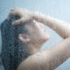 woman with dark hair taking a shower behind glass with her head up and hand on her forehead