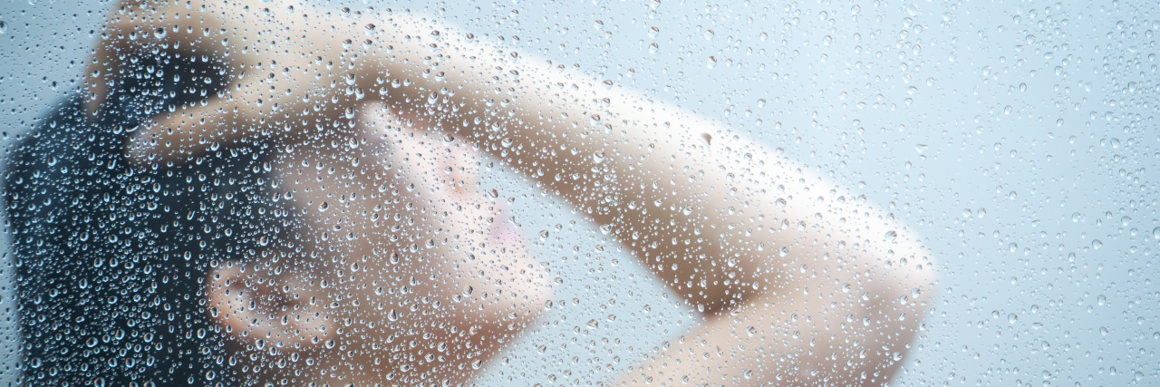 7 Helpful Showering Tips If Your Illness Makes Taking a Shower Hard woman with dark hair taking a shower behind glass with her head up and hand on her forehead