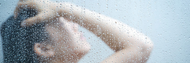 woman with dark hair taking a shower behind glass with her head up and hand on her forehead
