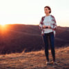 middle-aged woman in a flannel shirt and jeans standing on a hill during sunset looking out