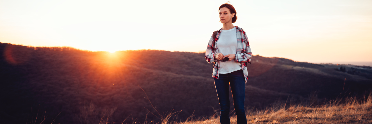 middle-aged woman in a flannel shirt and jeans standing on a hill during sunset looking out